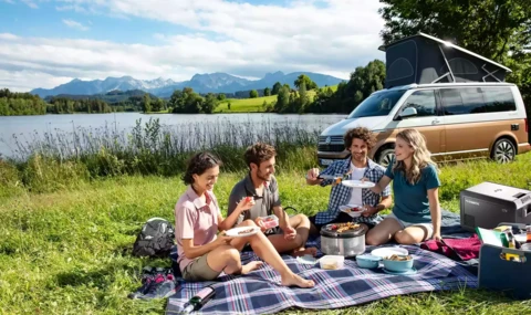 4 Personen sitzen auf einer Picknickdecke an einem See vor einem Campervan