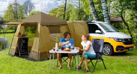 2 Personen sitzen an einem Picknicktisch mit Campingstühlen vor einem Zelt mit Campervan