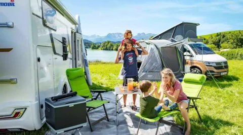 2 Erwachsene und 2 Kinder sitzen zwischen 2 Campervans auf einer Picknickdecke mit Tisch und Campingstühlen